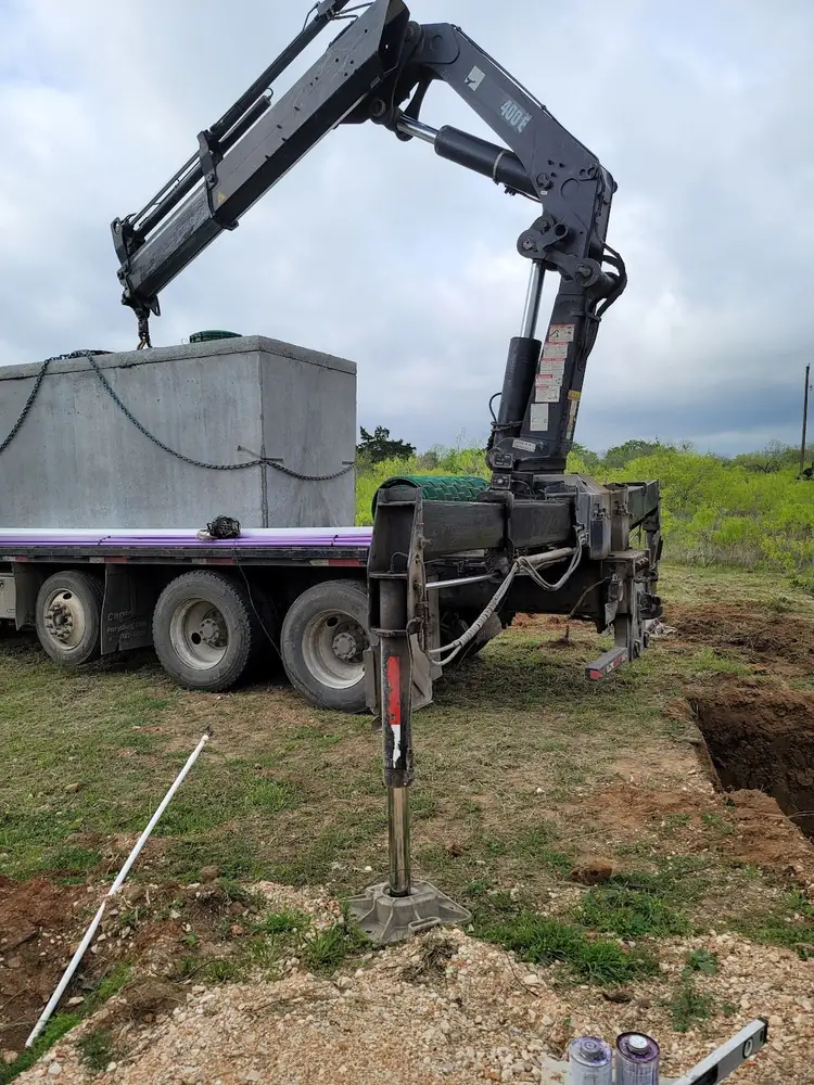 A boom truck lowering a concrete septic tank into place beside an open excavation