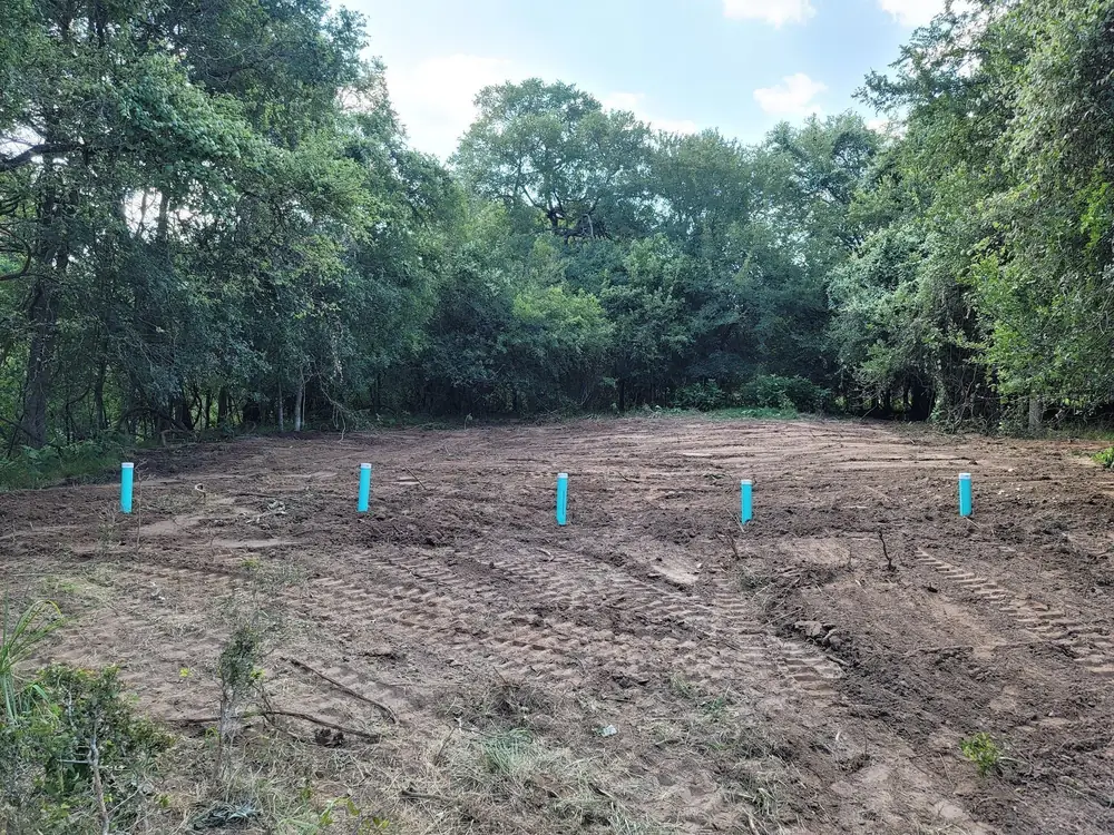 A newly graded drain field in a clearing ringed by trees, with blue marker pipes along the lines