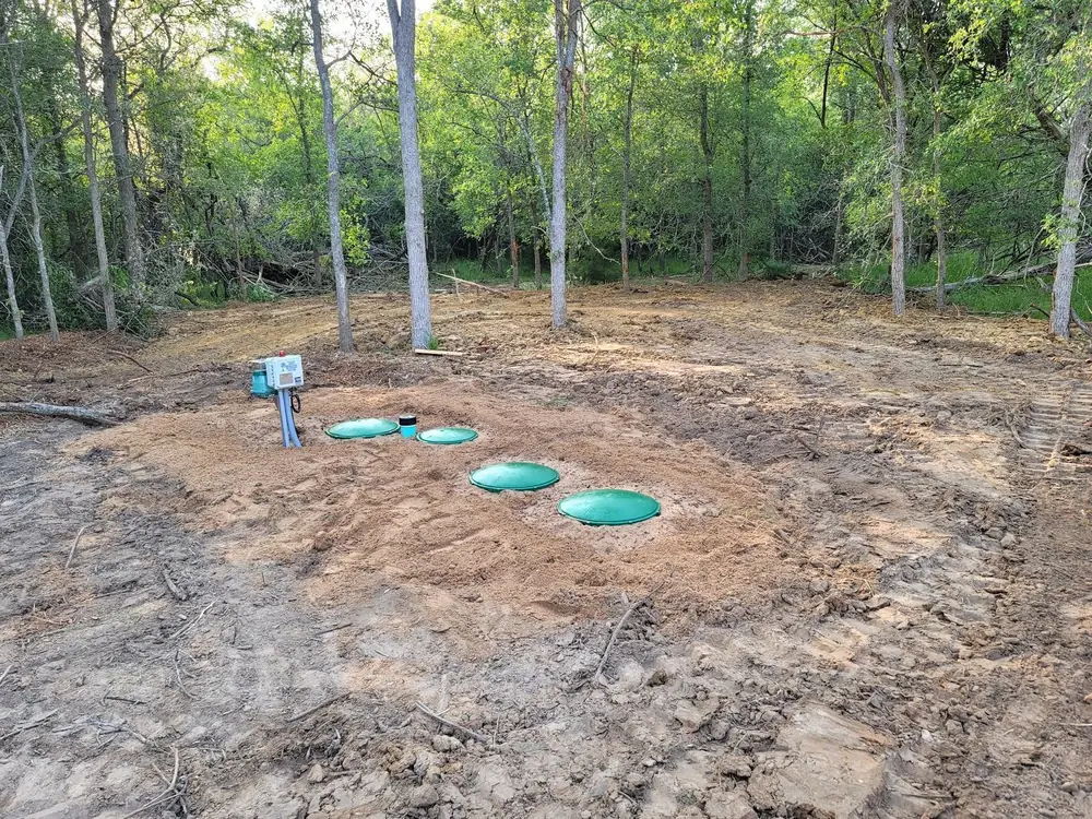 Finished aerobic system in a wooded lot: three green tank lids and a control panel in fresh sandy soil