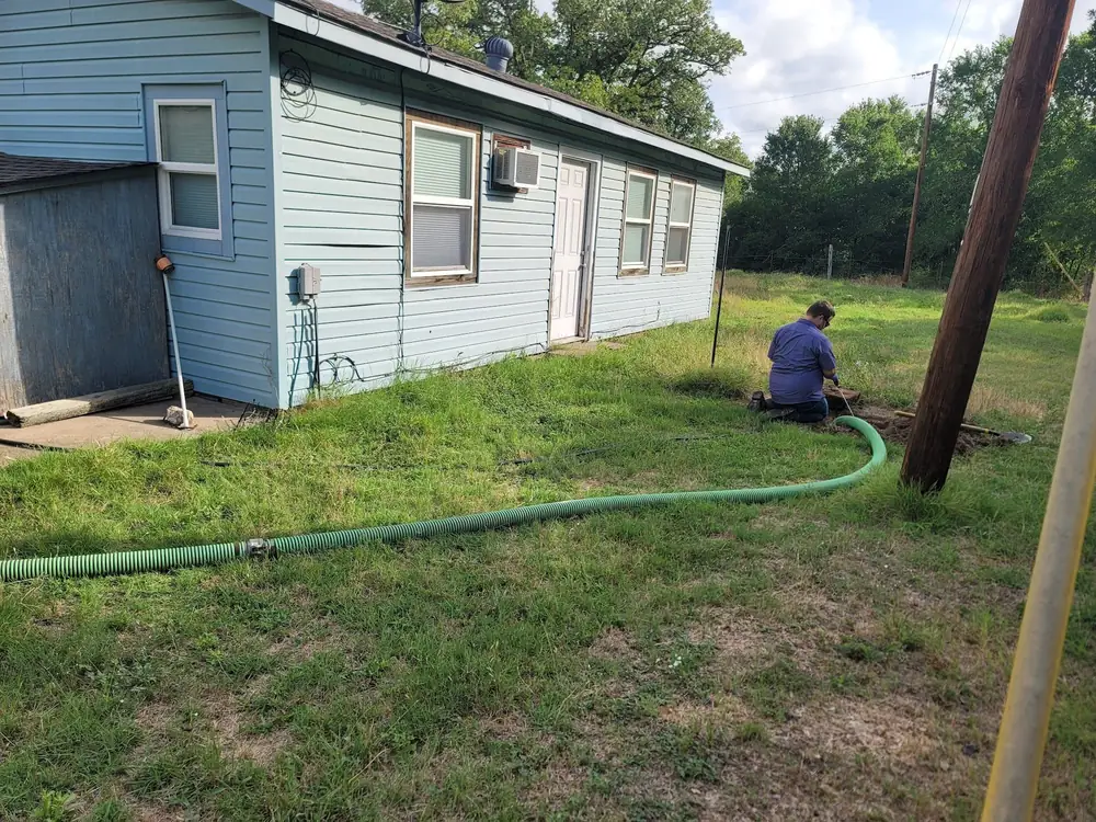 A technician pumping a septic tank beside a blue house, with the green vacuum hose across the lawn